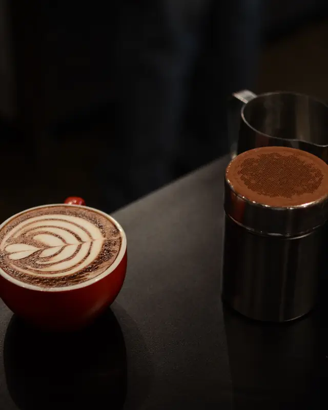 Eine rote Tasse mit Latte Art auf einem dunklen Tisch, daneben ein Milchkännchen.